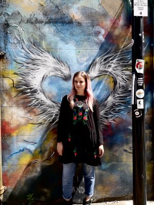 Pixie Tenenbaum standing in front of some street art angel wings in Shoreditch with a smaller street art caption in thre foreground which reads "Daddy I want a Fucking pony"